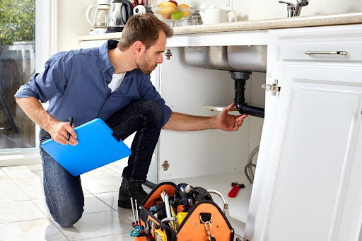 A plumber holding a blue clipboard and inspecting a black plumbing pipe in a kitchen in Carol Stream, IL.
