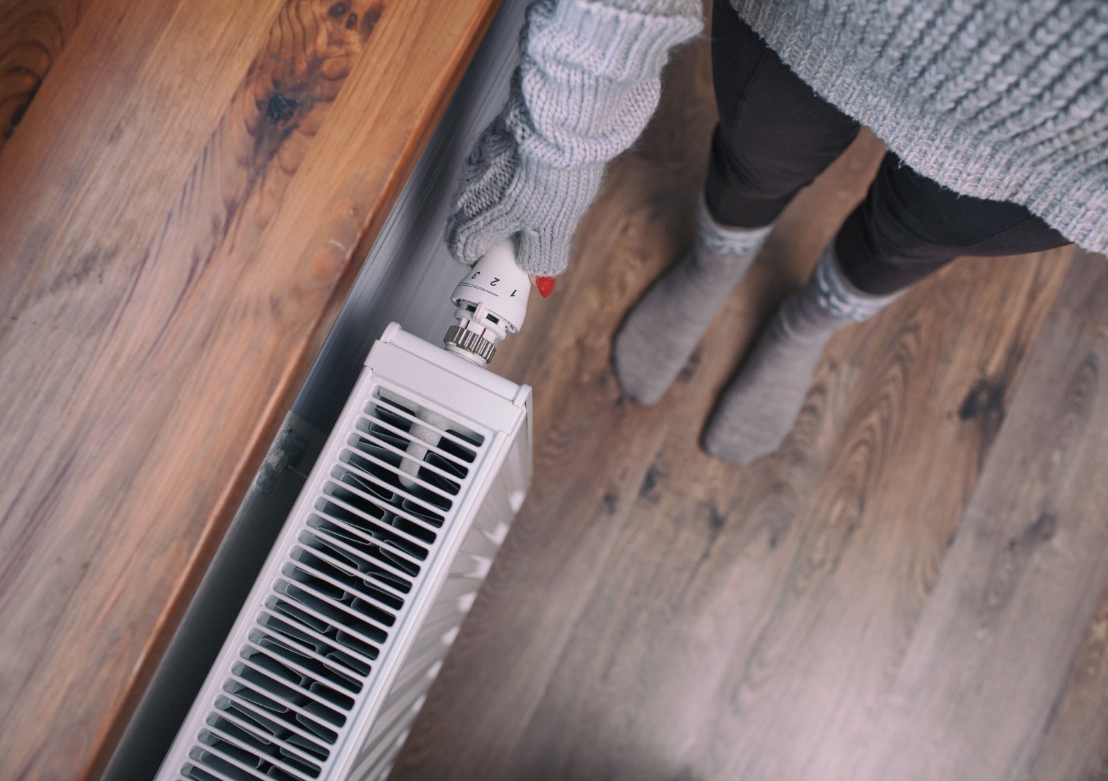 View from above of person wearing gray sweater, gloves, and tall socks, standing on wood floor while adjusting the temperature on a radiator.