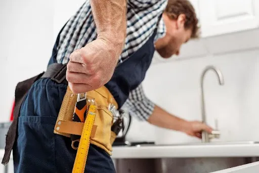 A plumber reaching for a tool in his work belt to repair a leaking kitchen faucet. An emergency plumbing service for a residential home.