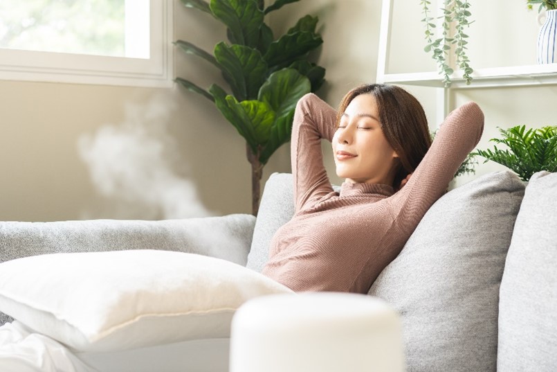 A modern air humidifier improves the indoor air quality and creates a comfortable living environment for a young woman relaxing on the couch with her arms behind her head in a home in Chicagoland.