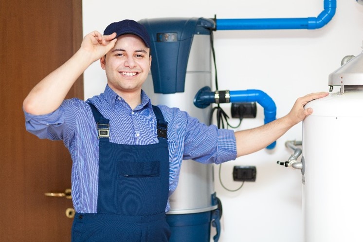 Smiling male plumber with one hand on his hat and one hand on a residential water heater in Carol Stream, IL.