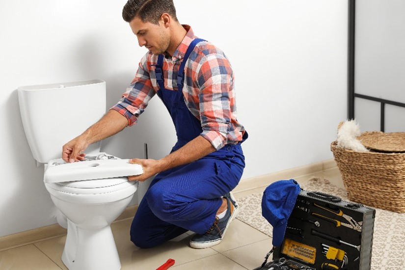 Professional male plumber with toolbox working on repairing a toilet in a bathroom in Chicagoland.