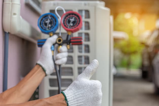 Close view of HVAC technician's gloved hands signaling a thumbs up and holding pressure gauge; AC system in background.