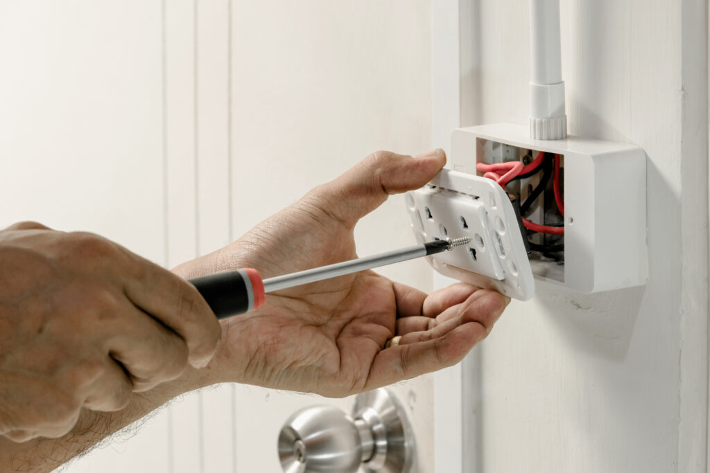 An electrician's hand using a screwdriver to attach an outlet cover to a wall outlet.