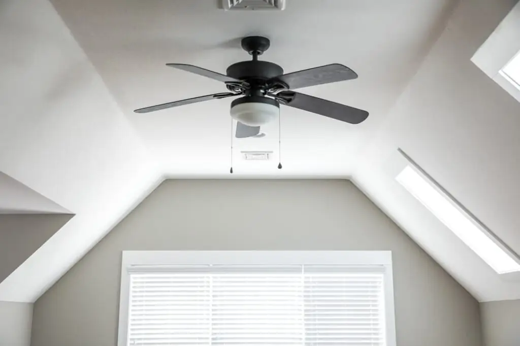 Open and airy bonus room in a new construction house with a dark wood ceiling fan, a window and blinds. White walls.